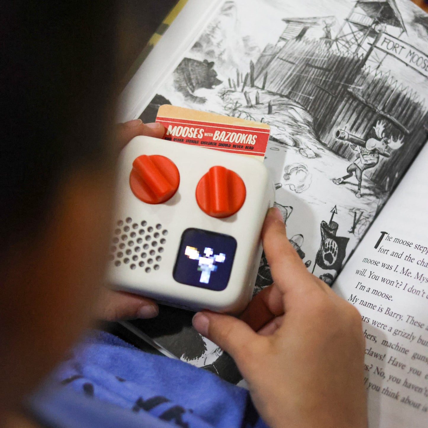 Person holding a Yoto device with red buttons and a screen, over a book with black and white illustrations.