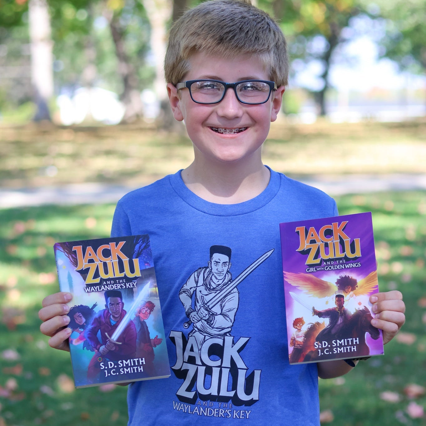 Young boy holding two books titled 'Jack Zulu' in a park setting